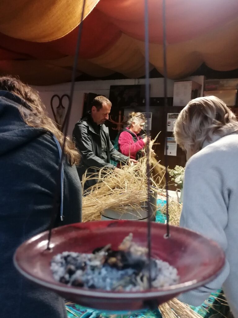 Räucherschale im Vordergrund, dahinter Menschen beim Basteln der Kornpuppe