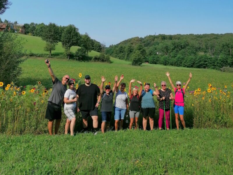 9 Personen stehend auf einer Wiese mit freudig erhobenen Armen, im Hintergrund eine Blumenwiese und hügelige grüne Landschaft