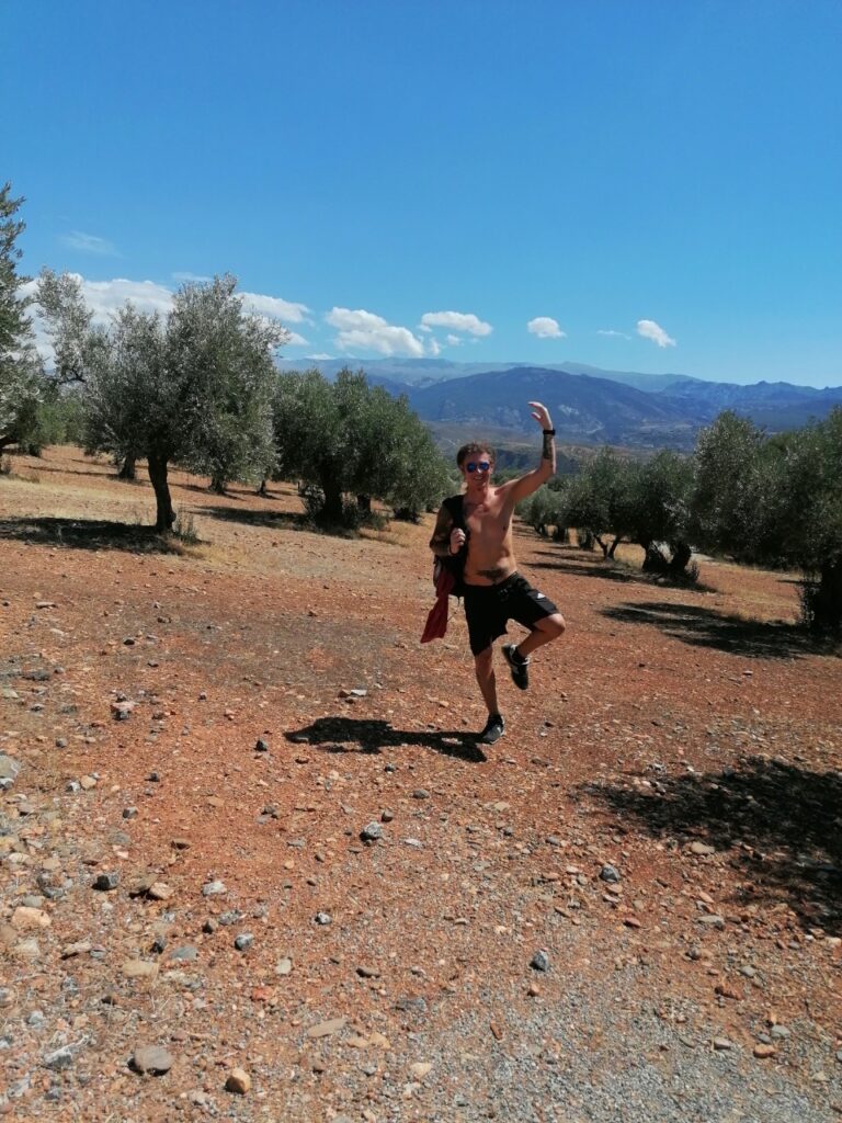 Seppi auf einem Bein auf roter, karger Erde - im Hintergrund Olivenbäume.