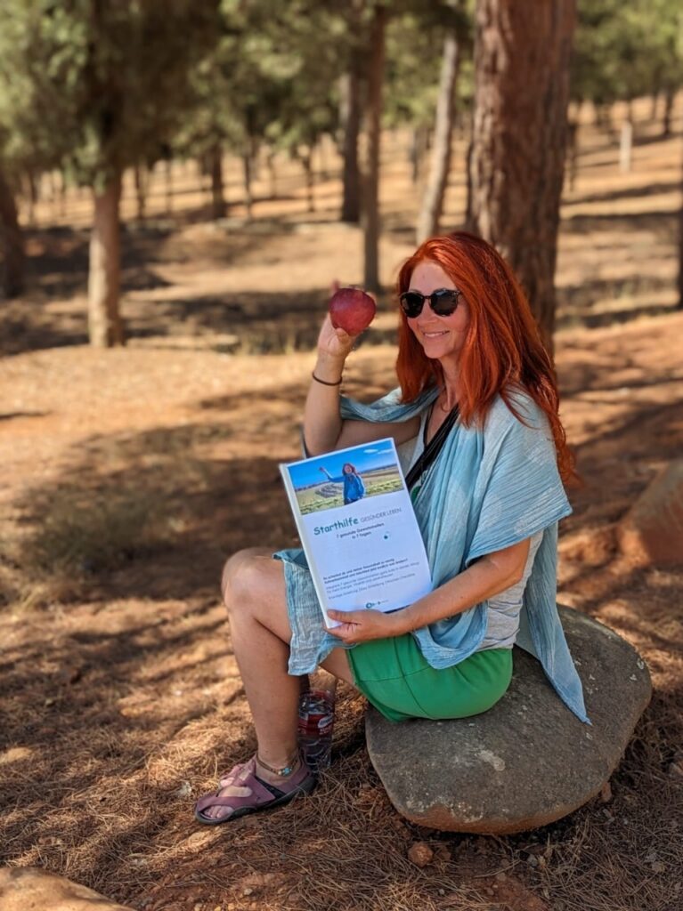 Karin mit Apfel und Starthilfe sitzend auf einem Stein, im Hintergrund rote Erde und Bäume