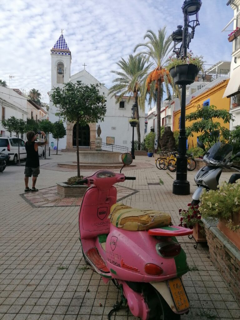 Straßenbild, im Vordergrund eine pinkfarbene Vespa, im Hintergrund eine Kirche