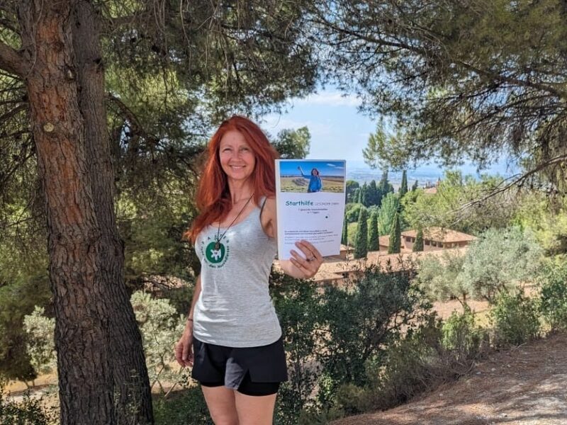 Karin Hartmann steht mit dem Ausdruck ihrer Freebie Starthilfe vor einem Baum. Weiter hinten sieht man klein die Alhambra.