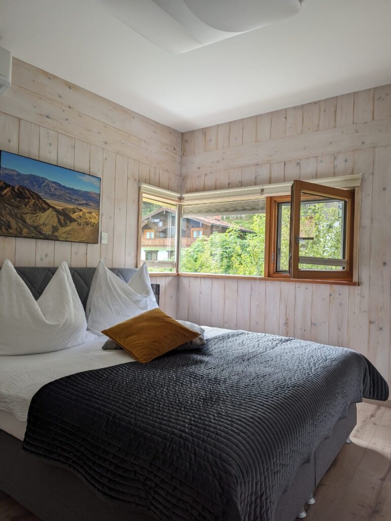 Doppelbett in einem hellen Zimmer mit schmalen Fenster und Blick ins Grüne