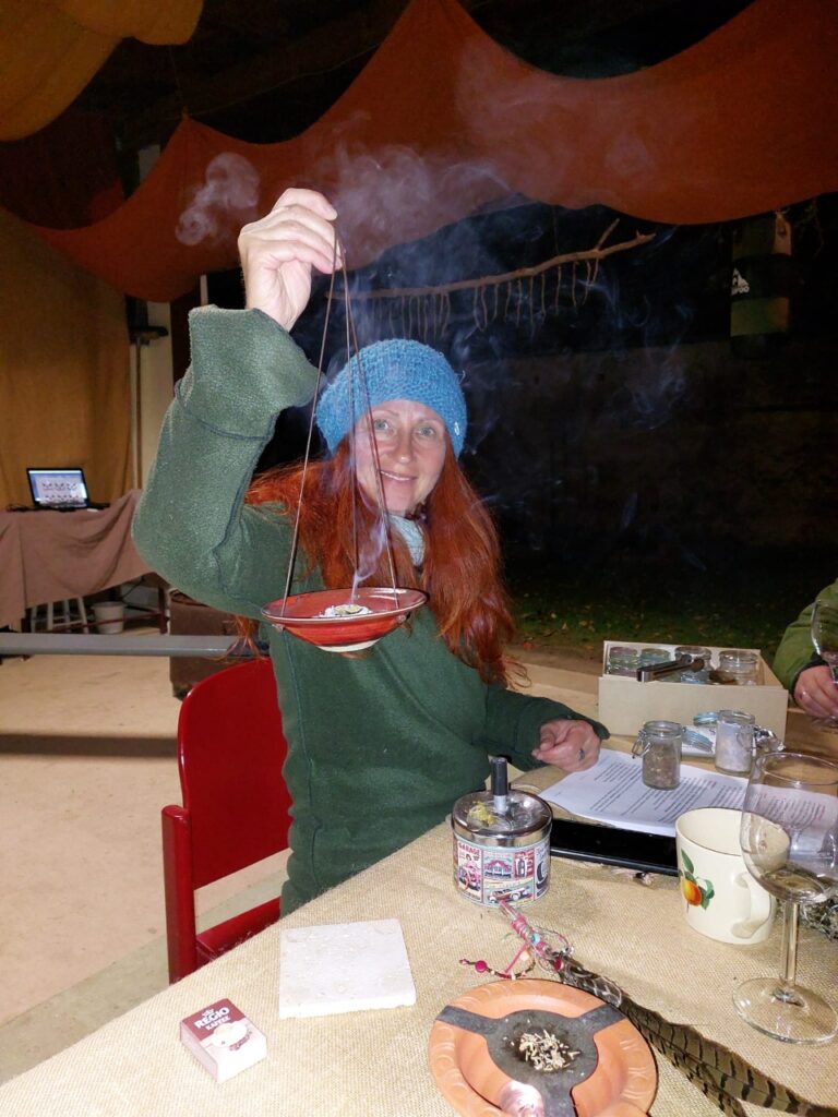 Karin mit grünem Mantel und blauer Haube, sitzend am Tisch mit Räucherschale in der Hand.