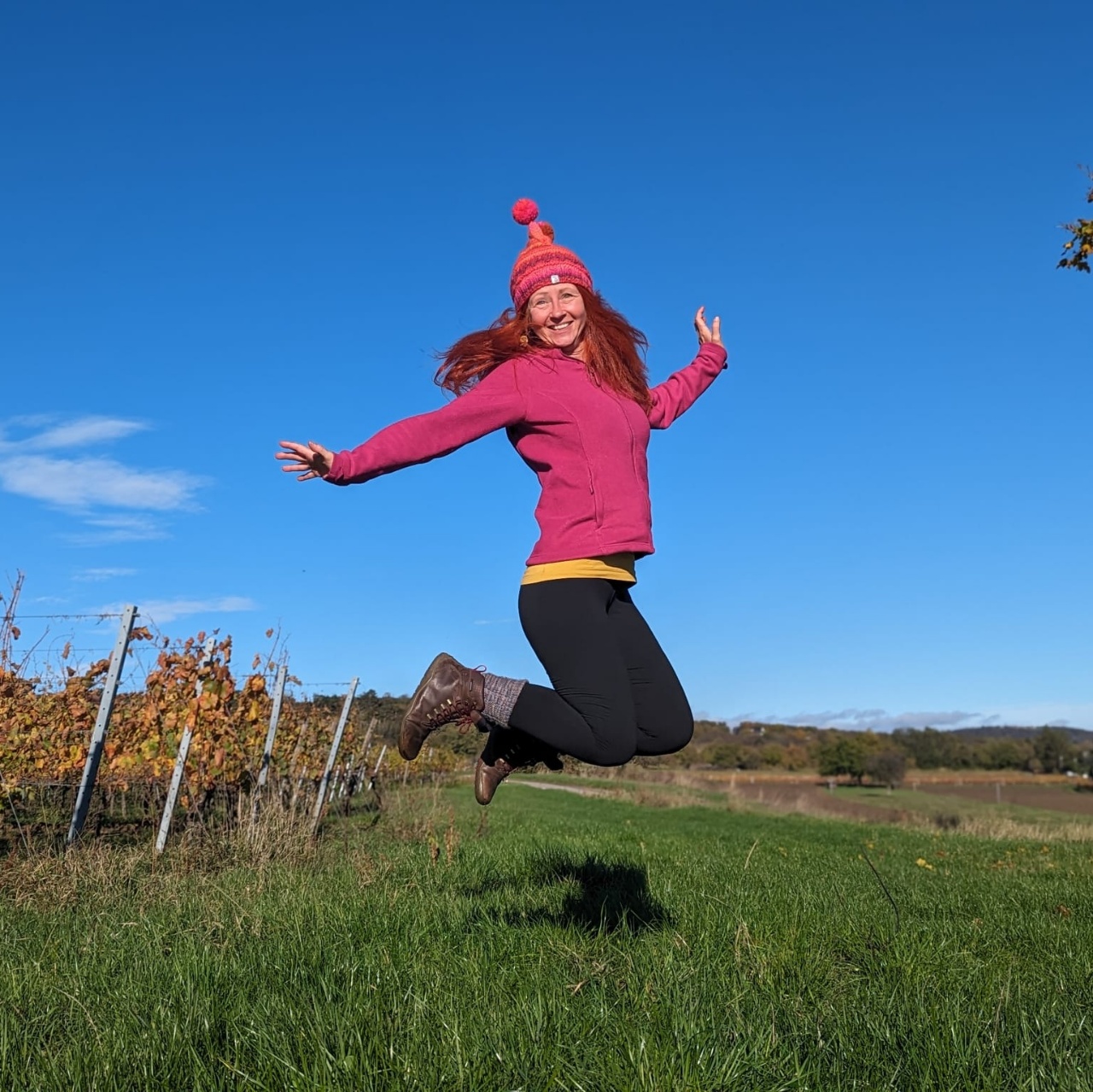 Karin macht einen Luftsprung. Sie hat eine pinke Mütze auf. Im Hintergrund blauer Himmel und Weingärten