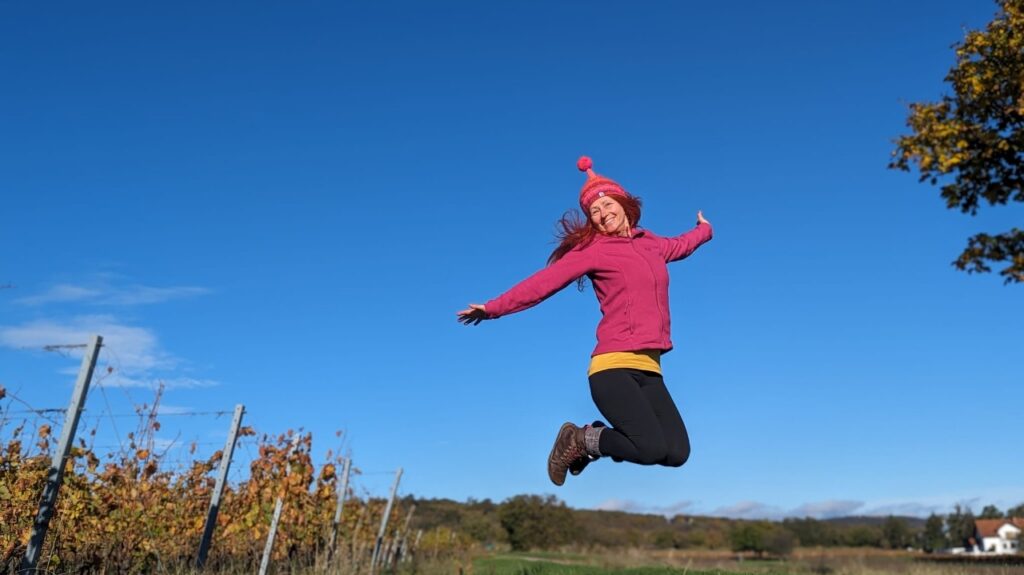 Karin im Sprung mit pinker Pudelhaube. Im Hintergrund blauer Himmel und Weingärten