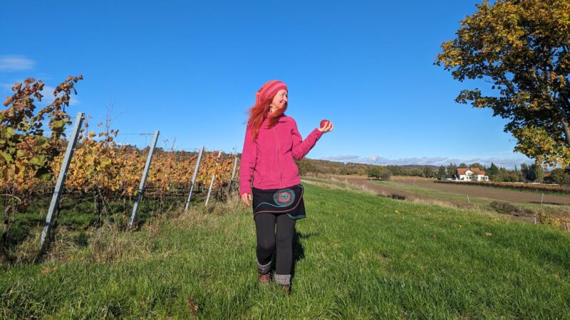 Karin mit pink Mütze und pink Weste und einen Apfel in der Hand, am Rande der Weingärten