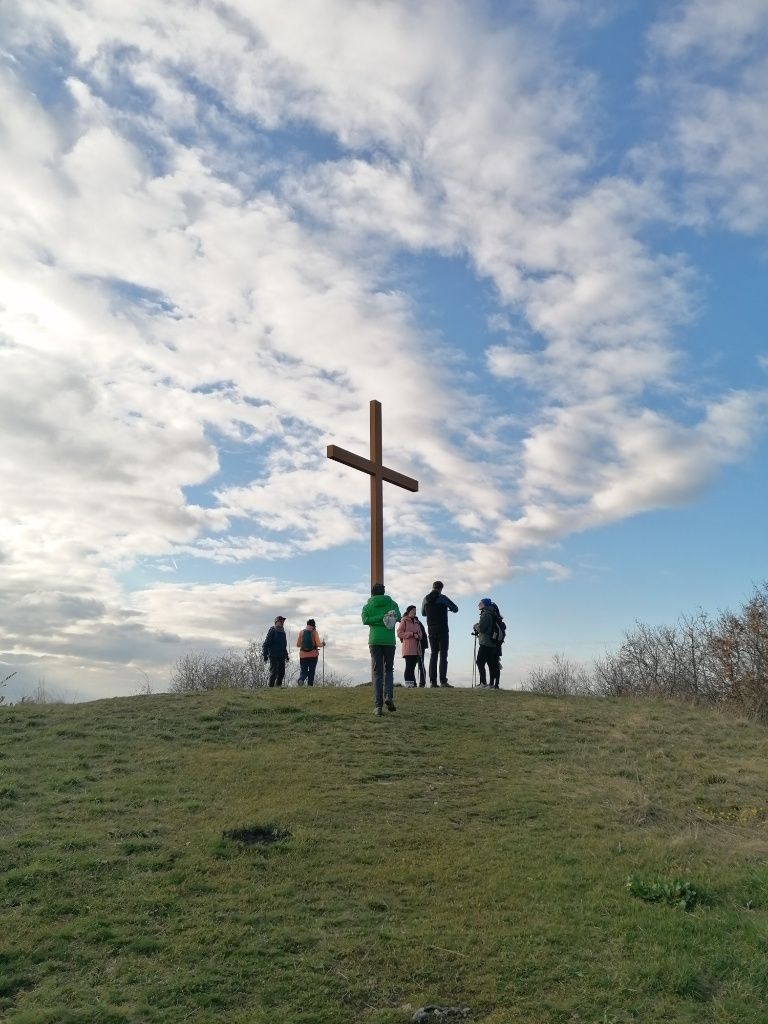 Gipfelkreuz mit 5 Personen aus der Ferne. Im Hintergrund bewölkter Himmel, im Vordergrund Wiese