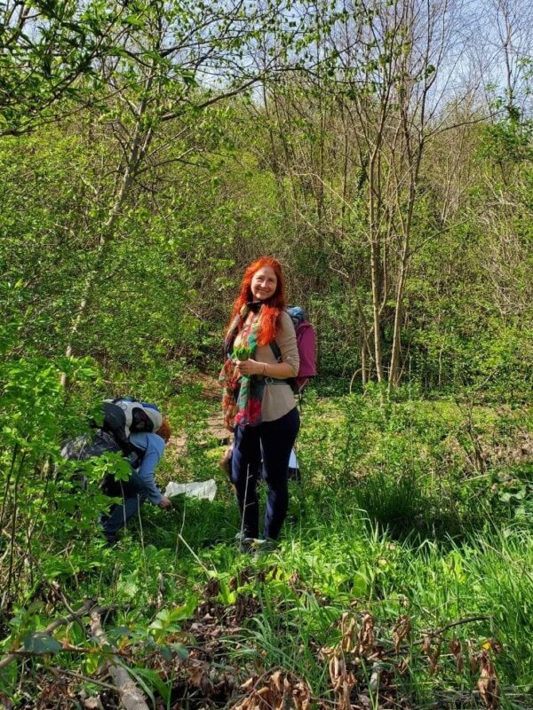Karin steht am Waldrand mit einem Buschen Bärlauchblätter in der Hand. Eva daneben in der Hocke beim Ernten.