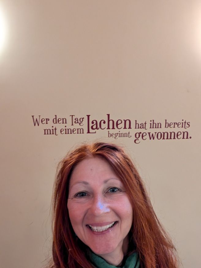 Karin vor einer Wand, auf der geschrieben steht: Wer den Tag mit einem Lachen beginnt, hat ihn bereits gewonnen.