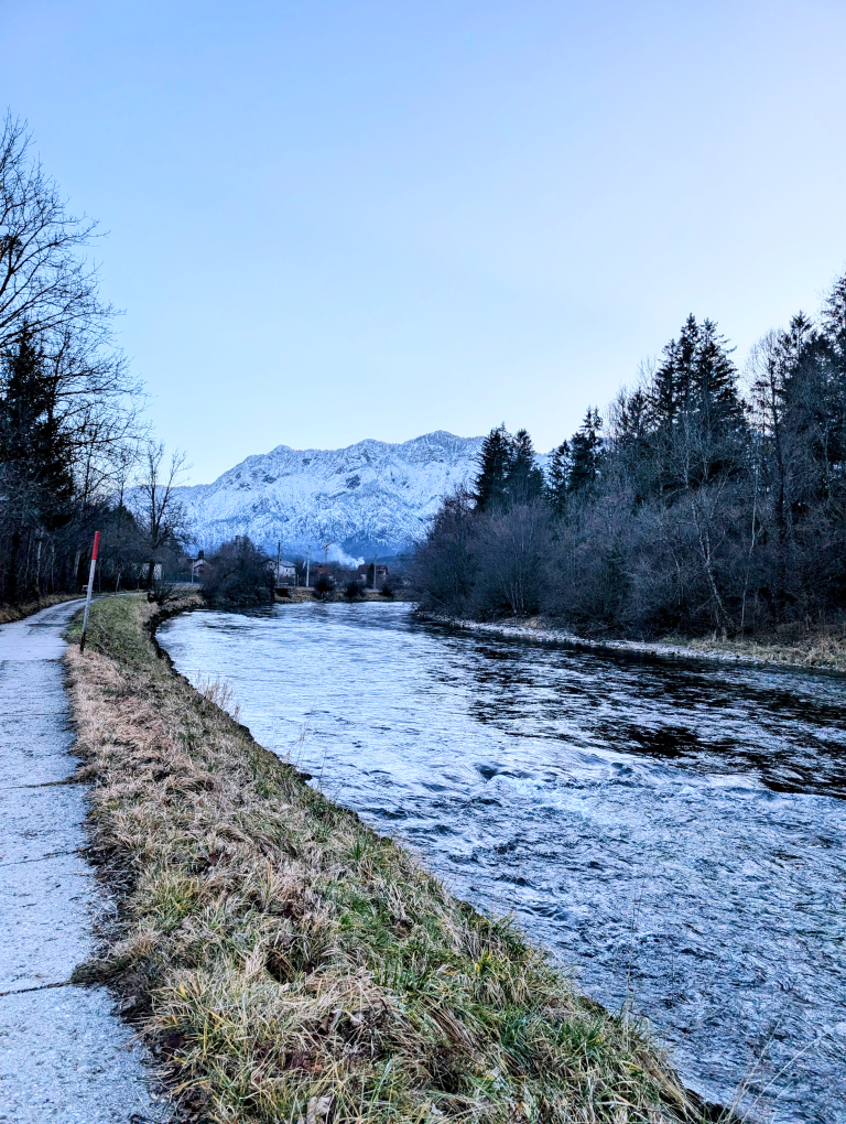 Fluß, links davon ein Weg, rechts Nadelbäume, in der Ferne Berge mit Schnee