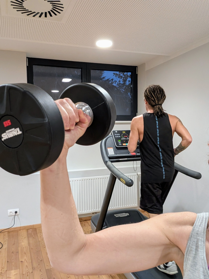 Karins Hand mit erhobener Hantel, dahinter Seppi auf dem Laufband.
