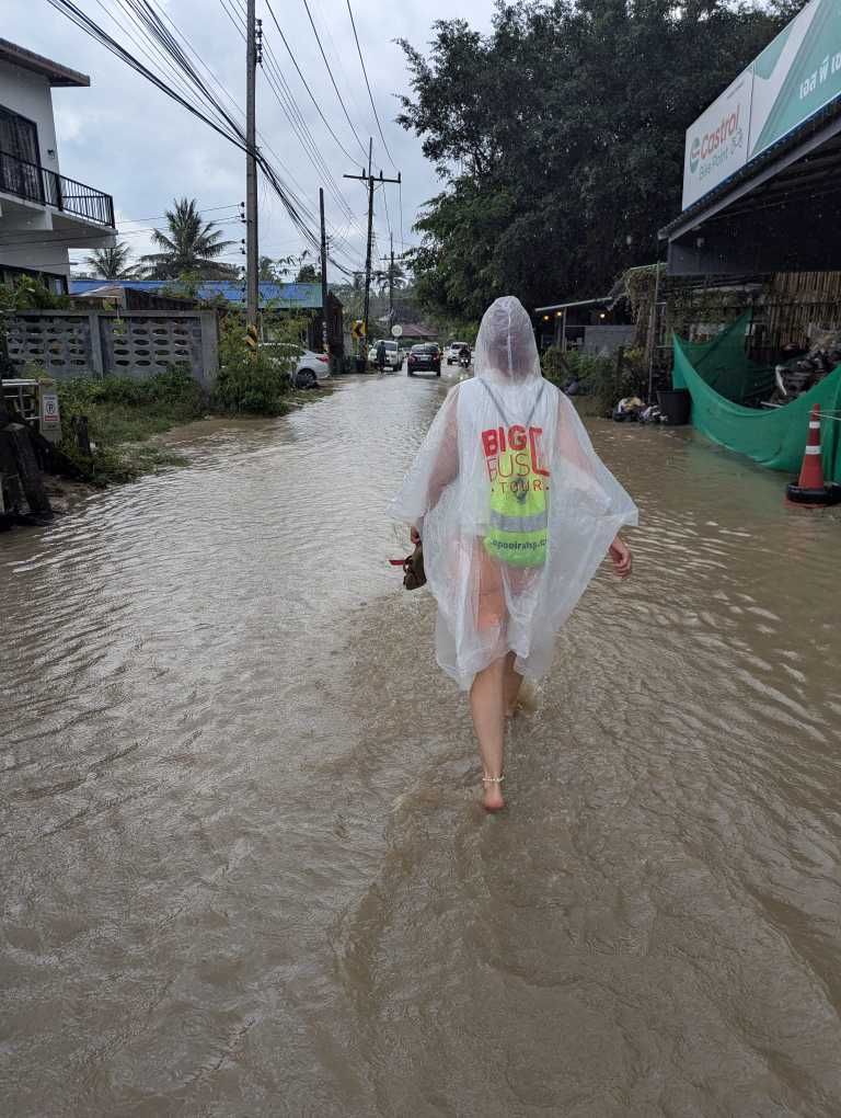 Karin barfuß mit Regencape und Sandalen in der Hand durch Riesenpfütze hindurch marschierend