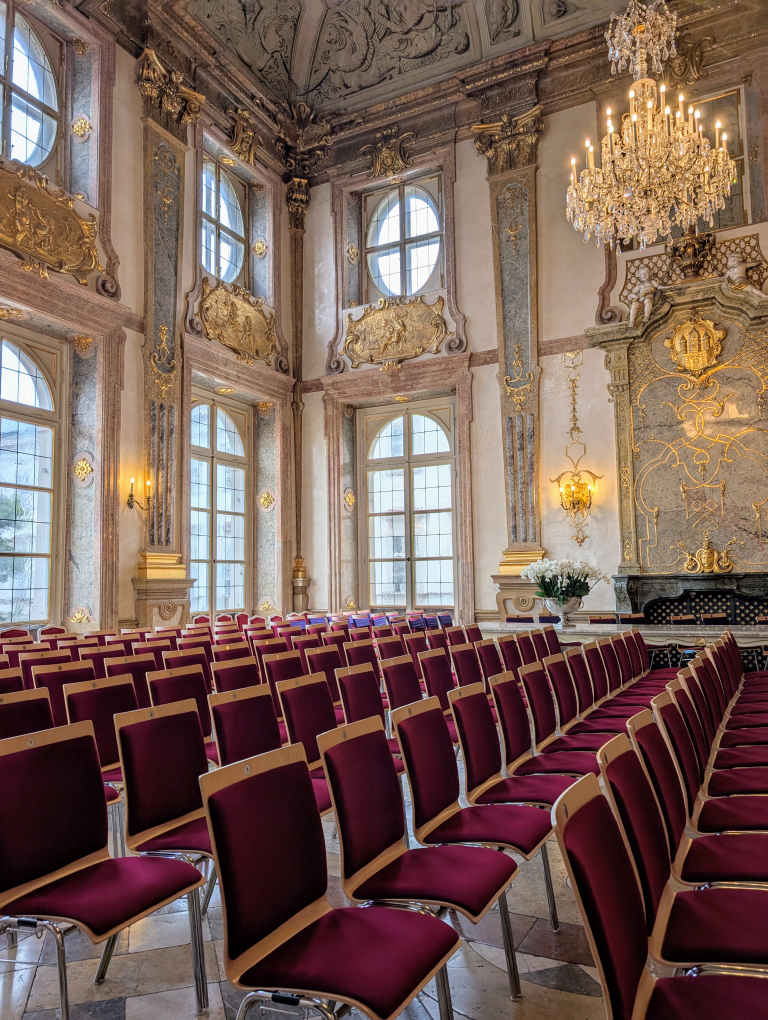 Rote Sesselreihen in einem Saal mit hohen, verschnörkselten Fenstern und Kronleuchtern