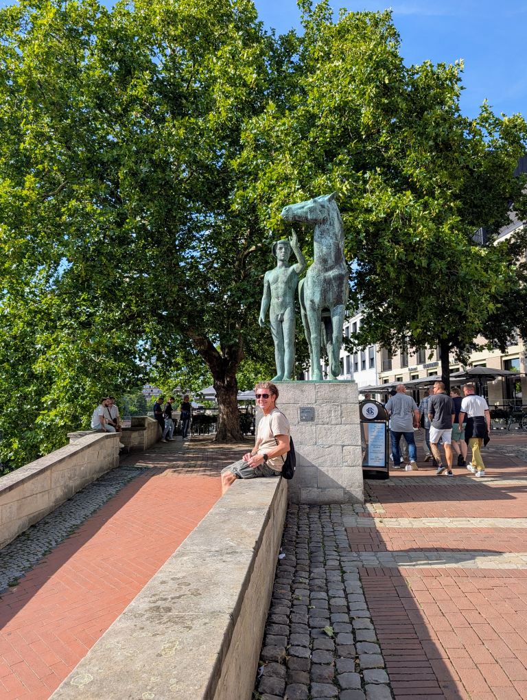 Seppi sitzt auf einer Betonmauer, im Hintergrund eine Pferdeskulptur mir einem Mann
