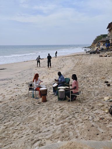 Karin mit Oumar und seinem Sohn beim Trommeln am Strand, im Hintergrund tanzen zwei Männer