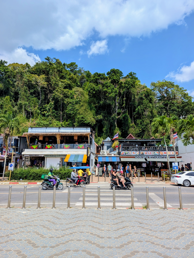Zwei traditionell thailändische Shops von der gegenüberliegenden Straßenseite. Hinter den Gebäuden ist ein Meer aus grünen Bäumen.
