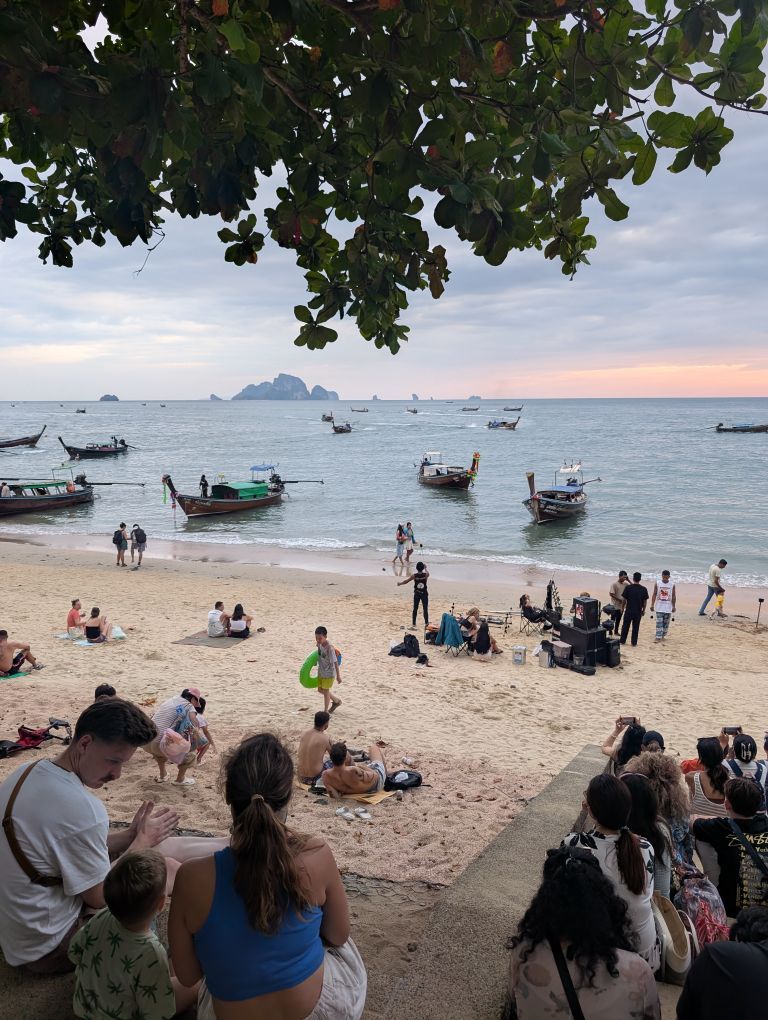 Strand in den Abendstunden mit Menschen und Booten. In der ferne kleine Felsinseln.