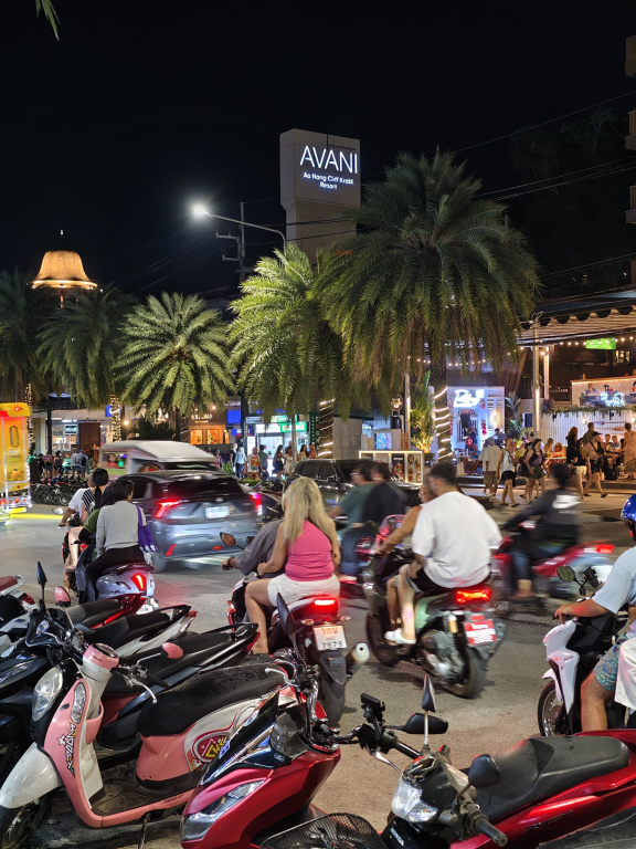 Belebte Straße bei Nacht, mit vielen Mopeds, Autos und Palmen am Straßenrand.