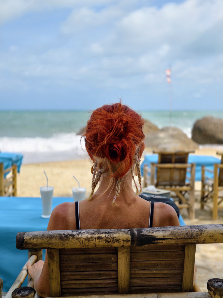 Karin von hinten mit Blick aufs Meer, Die langen roten Haare sind nach oben gesteckt, die kurzen, nachgewachsenen grauen Haare im Nacken sind zu kleinen Zöpfchen geflochten. Schaut cool aus. Am Tisch vor Karin stehen zwei Bananen Shakes
