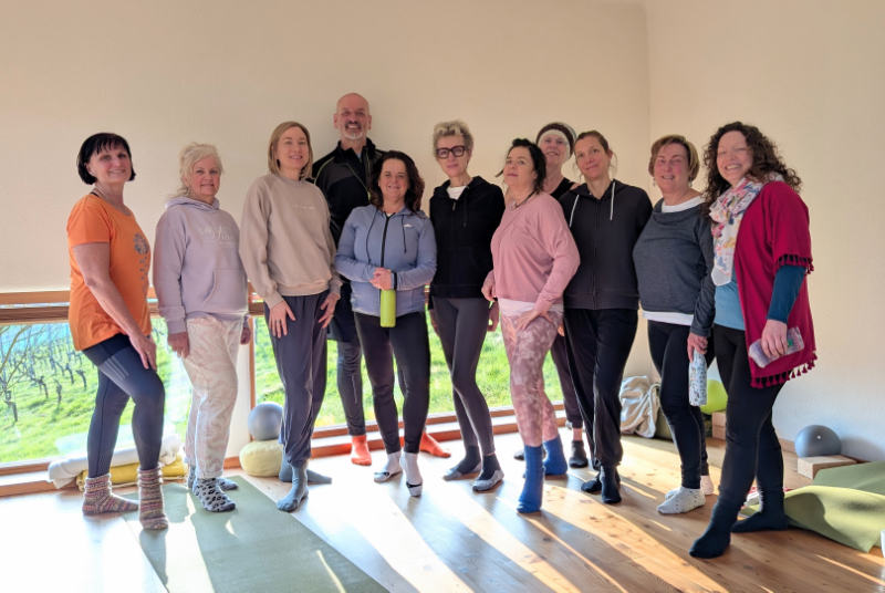10 Frauen und 1 Mann im Yoga-Outfit, stehend im sonnendurchfluteten Yoga-Raum
