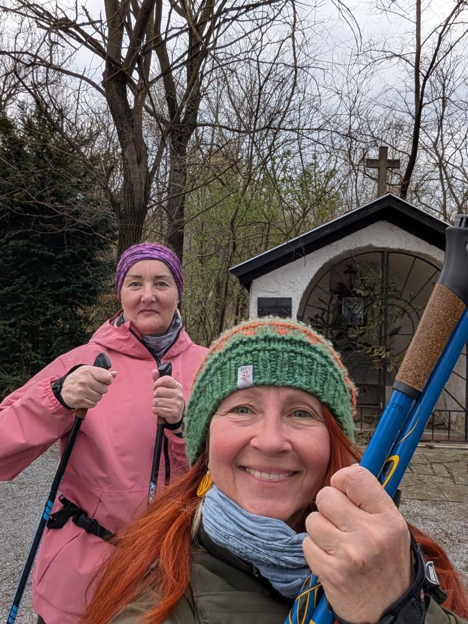 Karin und Anita mit erhobenen Nordic Walking Stöcken vor einer Kapelle im Wald.